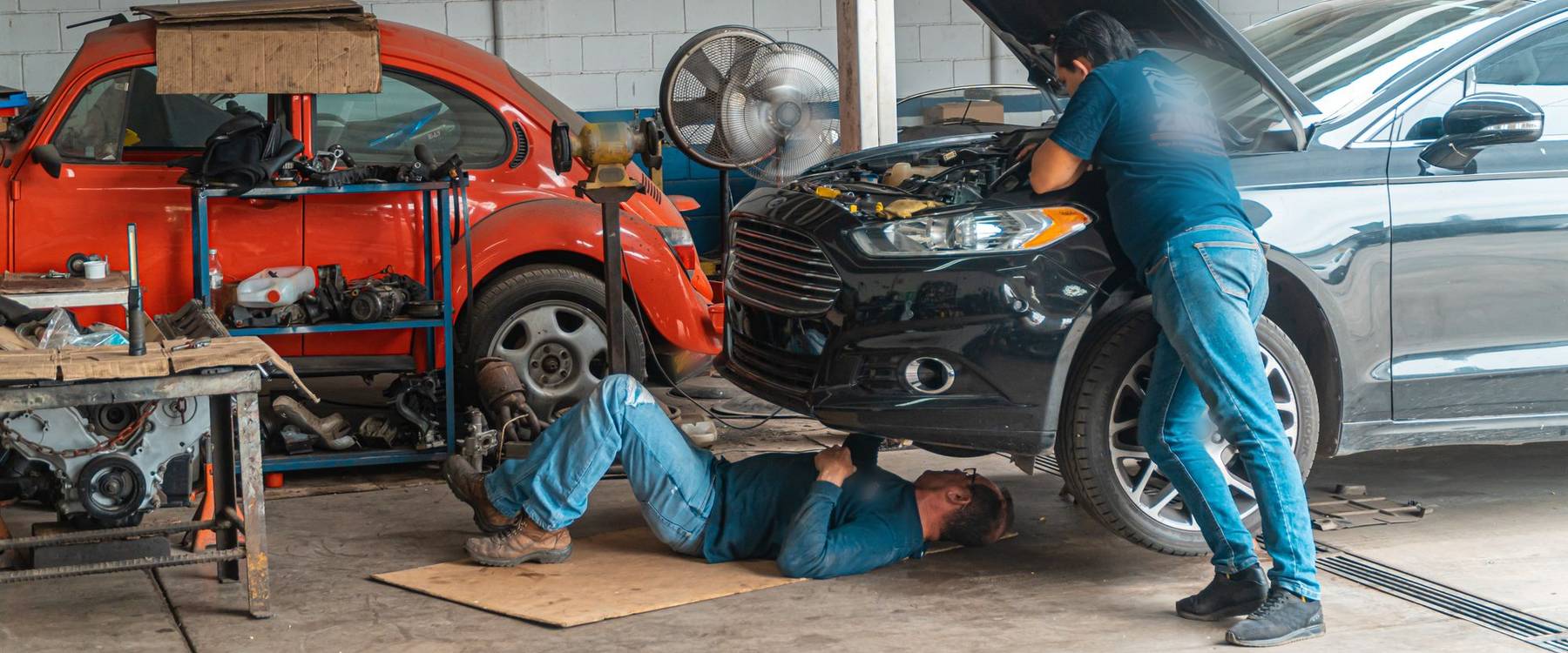 Dois mecânicos numa oficina, um deitado sob um sedã preto e outro inclinado sobre o motor com o capô aberto
