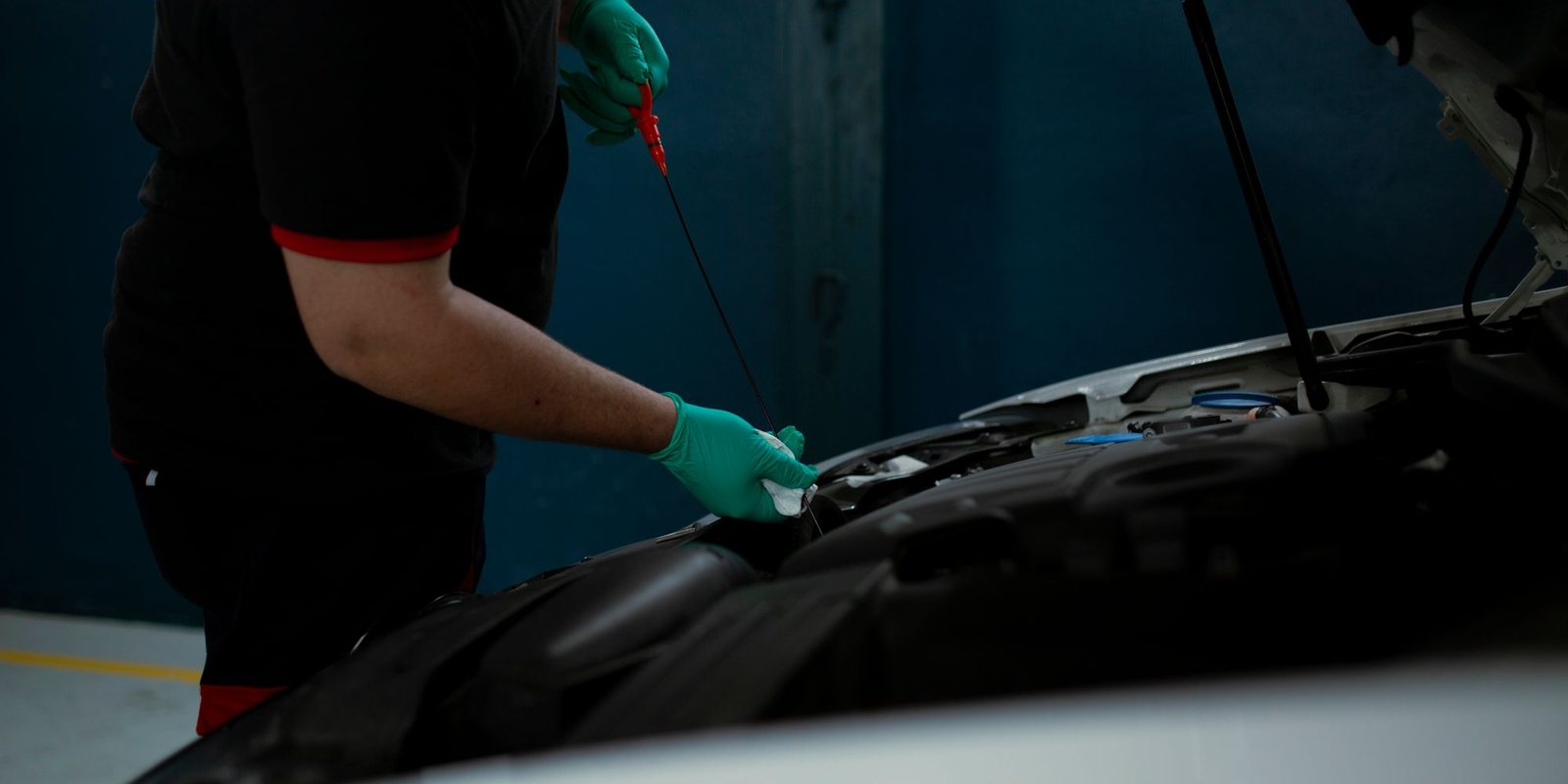 Mão com luva verde limpa a vareta de medir o nível do óleo diante do motor aberto